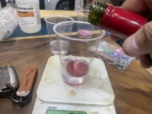 透明レジンの入った計量カップに赤色の着色剤を少量ずつ加えている様子。レジンの色づくり工程。