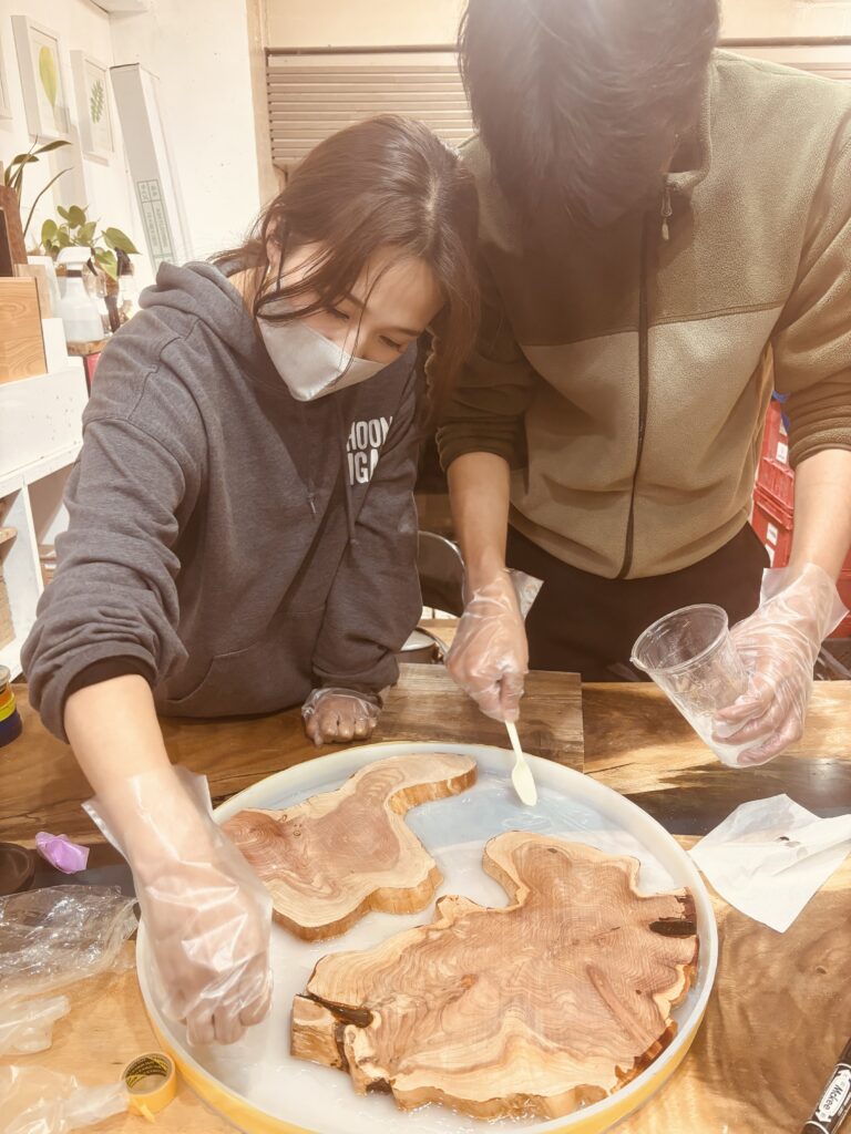 レジンテーブル制作体験に参加した夫婦が並んで作業し、木材の位置決めとレジン塗布を行っている様子。