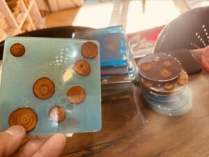 木の輪切りパーツを封入したカラフルなレジンコースターを手に持ち、積み重ねられた試作品が背景に映っている様子。