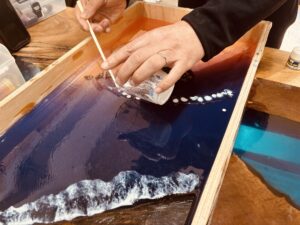 レジンテーブル制作体験で、福井県雨晴海岸の夕景をイメージしたブルーの海に白い波のレジンを流し込む工程