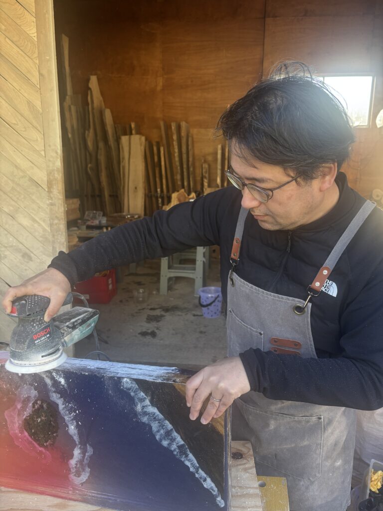 レジンテーブル制作体験で、受講者が雨晴海岸の夕景を表現したレジン天板をオービタルサンダーで研磨している様子