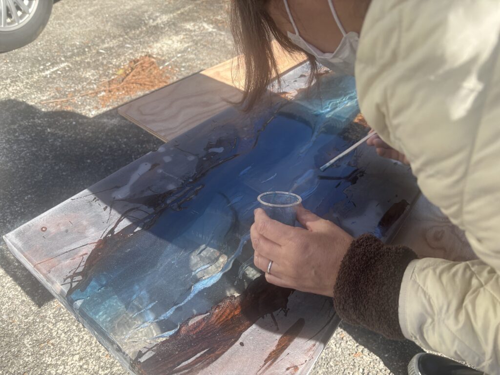 レジンテーブル制作体験で、ブルーからクリアへのグラデーションを筆で描いている工程。カップに入ったレジンを使い、天板上で細かな調整を行っている様子。